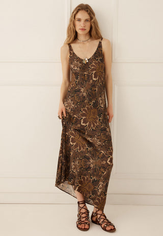 A woman modeling a sleeveless brown maxi dress with a metallic bronze floral pattern, paired with brown strappy sandals, standing against a white paneled wall.