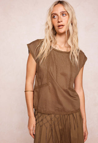 A woman with long blonde hair wearing a brownish-olive brown sleeveless top and matching bottoms, standing against a textured light beige wall.