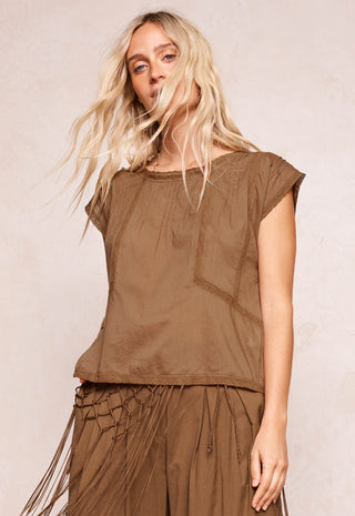 A woman with long blonde hair wearing a brown short-sleeve top and matching brown fringe skirt, posing against a neutral beige background.