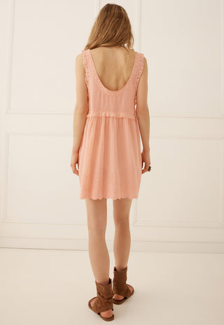 A woman viewed from behind wearing a sleeveless peach-pink mini dress with a ruffled scoop neckline and brown leather slide sandals, standing against a white paneled wall.